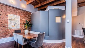 A dining area with a brick wall, a modern gray accent wall, wooden ceiling beams, and a dining table with chairs.