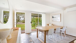 A modern dining area with a wooden table and white chairs, a large window with a view of a garden, and a framed picture on the wall. - Hanalei - Pet Friendly - Directly Opposite Waterfront (Myola)