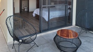 Een moderne buitenruimte met een schuifpui naar een slaapkamer, twee zwarte draadframe stoelen en een houten bijzettafeltje.
