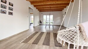 Een ruime kamer met een houten plafond, een hangmat, een eettafel met stoelen en uitzicht op de omgeving door een groot raam.