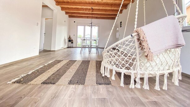 Een ruime kamer met een hoog houten plafond, een hangstoel van macramé en een eethoek op de achtergrond.