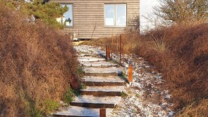 A modern building with a wooden exterior and large windows, accessed by a stone staircase surrounded by grass and bushes. - Impeccable Studio in Ouddorp 400m from th (Ouddorp)
