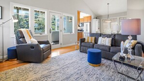 A spacious living room with a gray sofa, blue ottoman, and a glass coffee table. Large windows offer a view of a hillside.