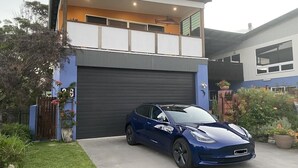 Una casa moderna de dos pisos con garaje, un coche azul estacionado al frente y un jardín bien cuidado.
