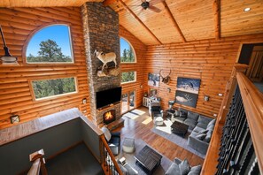 A log cabin interior with a stone fireplace, wooden walls, a ceiling fan, and a large window overlooking the outdoors.