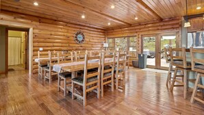 A wooden cabin with a dining area featuring multiple wooden tables and chairs, a clock on the wall, and a view through the glass doors to the outside.