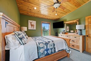 A bedroom with a wooden ceiling, a bed with a patterned blanket, a dresser with a mirror, and a television on the wall.