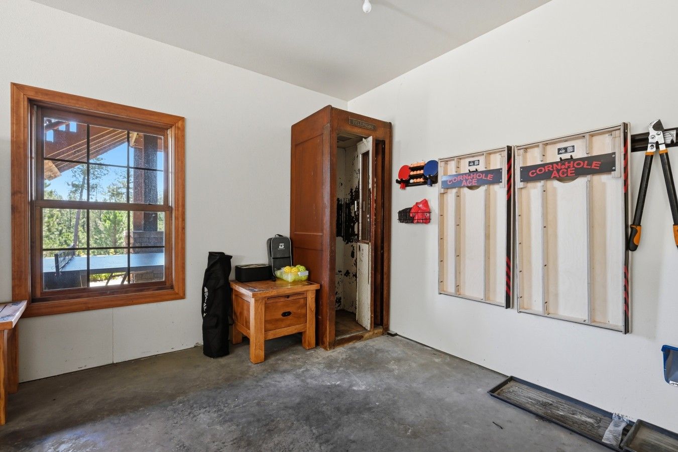 A room with a large window, a wooden desk, a small table with a bag, and a wall with signs reading 'CORN-HOLE ACE'.