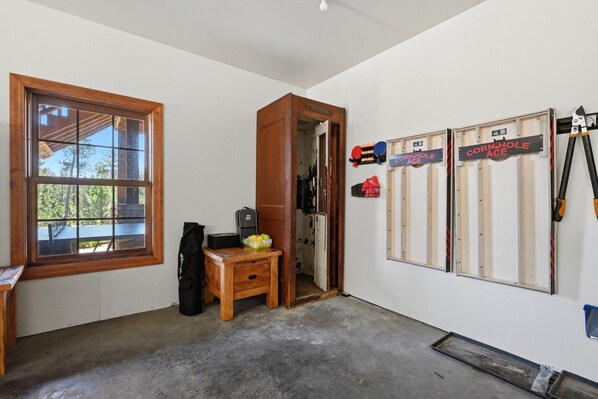A room with a large window, a wooden desk, a small table with a bag, and a wall with signs reading 'CORN-HOLE ACE'.