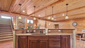 A log cabin interior with a central island kitchen, wooden ceiling, and a stone fireplace.