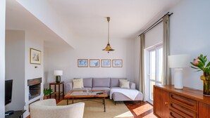 Ein Wohnzimmer mit einer Couch, Couchtisch und Fernseher. An den Wänden hängen Bilderrahmen und ein Fenster mit Vorhängen.