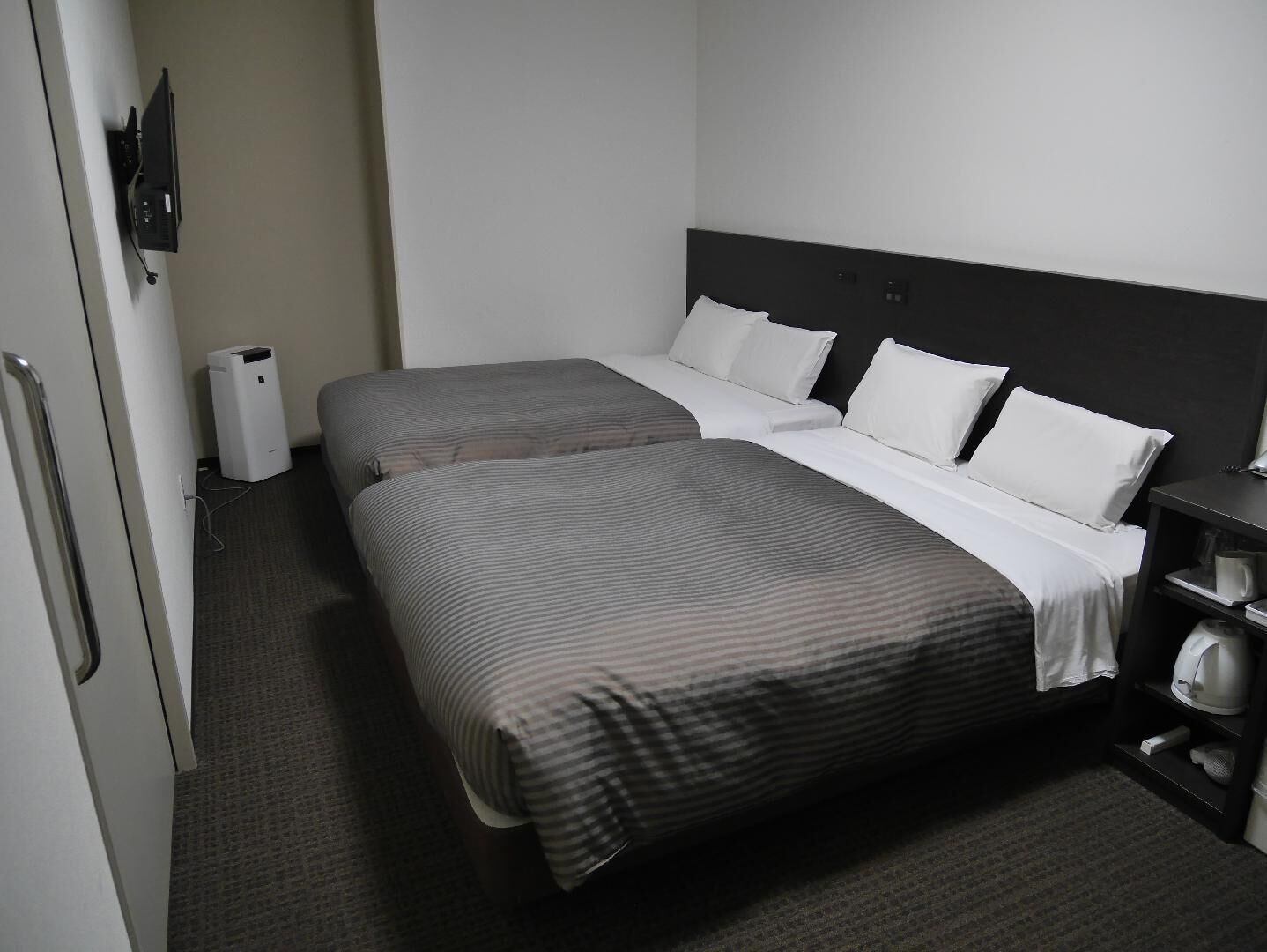 A hotel room with two beds, a bedside table, a lamp, and a small shelf with a kettle.