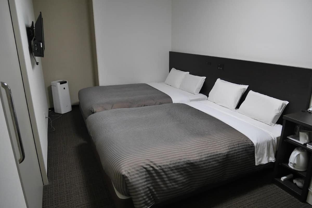 A hotel room with two beds, a bedside table, a lamp, and a small shelf with a kettle.