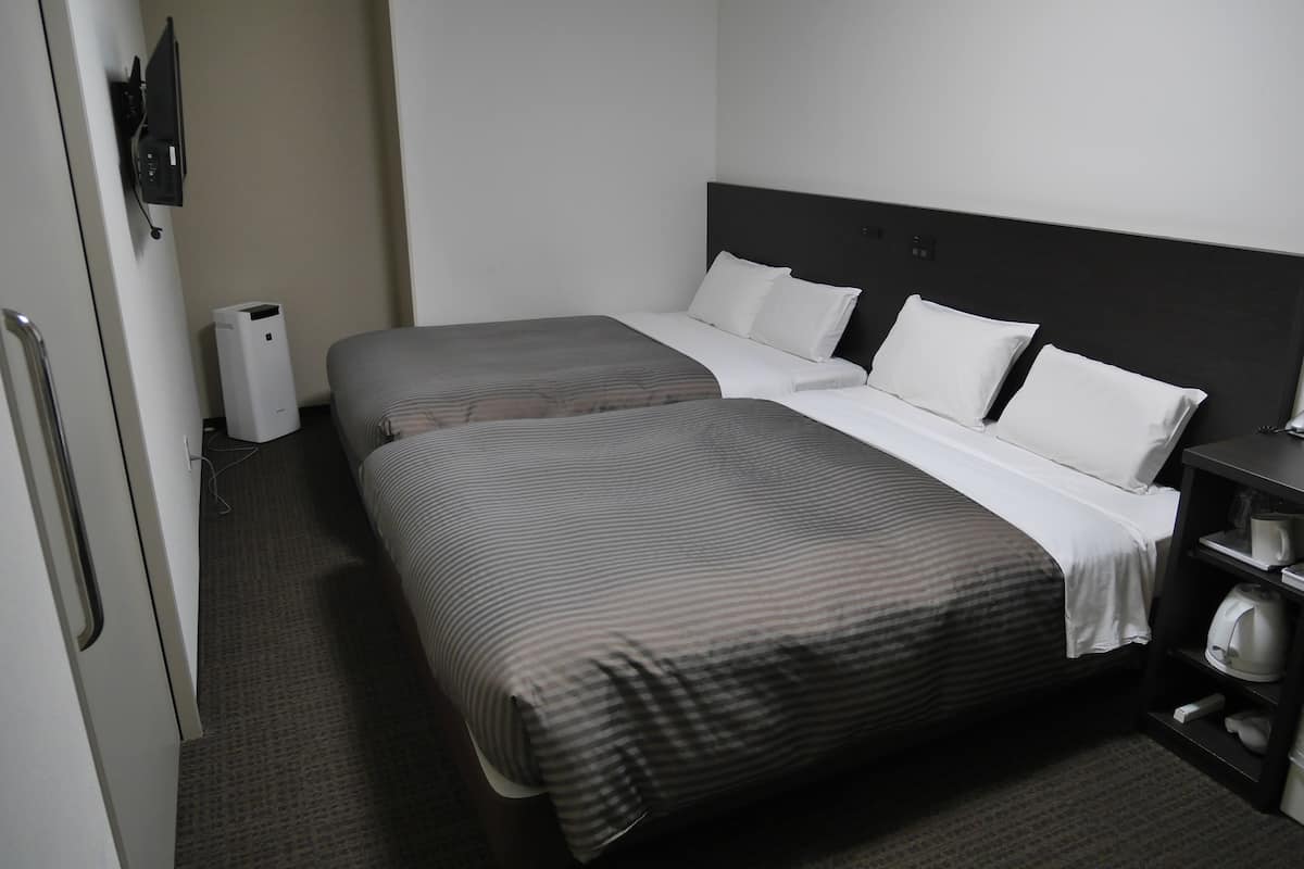 Quarto de hotel com duas camas, mesa de cabeceira, abajur, aquecedor e guarda-roupa.
