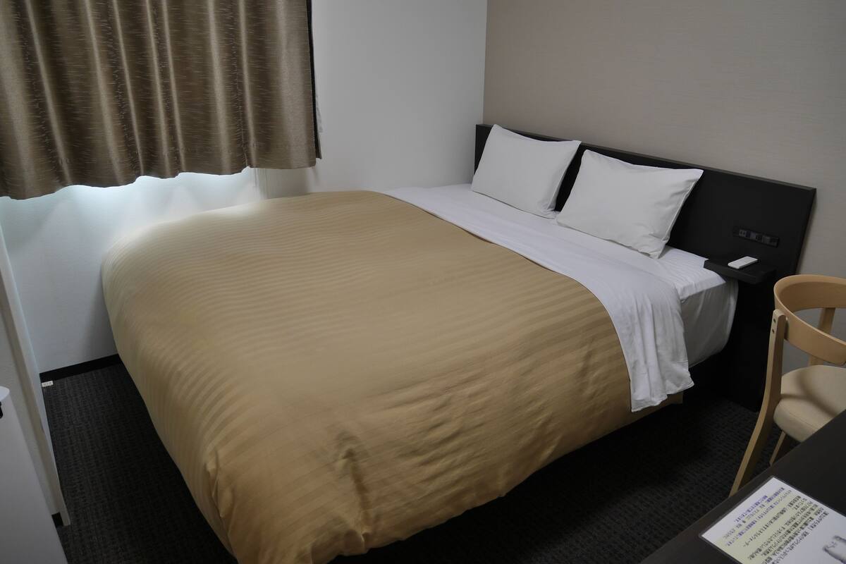 Quarto de hotel com cama, uma cadeira, mesa de cabeceira e janela com cortinas.