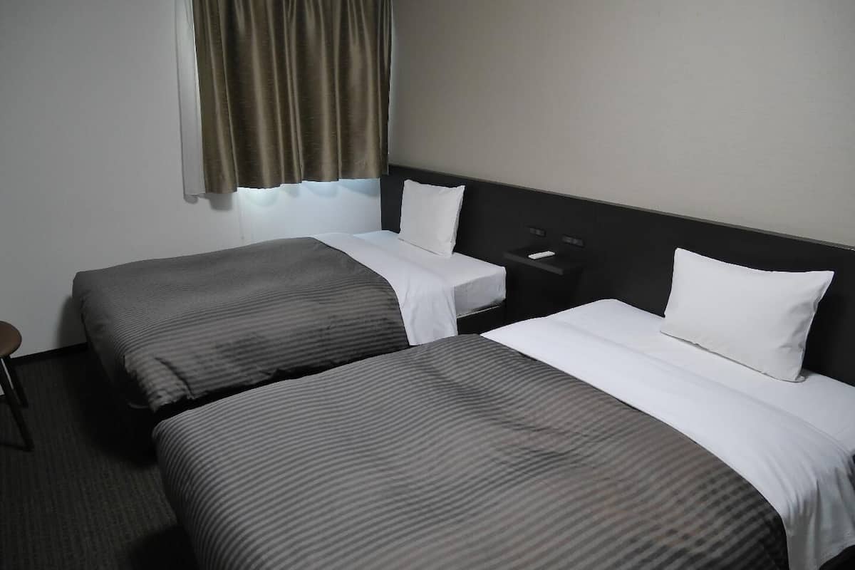 Quarto de hotel com duas camas, uma mesa de cabeceira, uma cadeira e uma janela com cortinas.