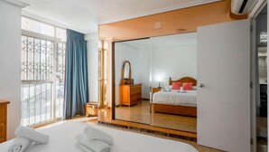 A hotel room with a large bed, a wooden dresser, a mirror, and a view through a window with blue curtains. - Rinconada Real by Interhome (Benidorm)