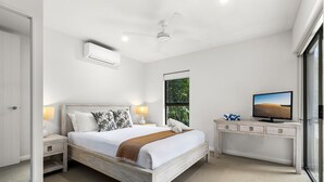 A bedroom with a bed, nightstands, a desk, a television, and a window with a view of greenery.