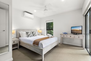 Un dormitorio con cama, mesitas de noche, escritorio, televisor y ventana con vista a zonas verdes.
