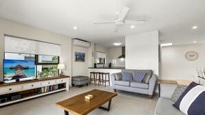 A modern living room with a TV, sofa, coffee table, and a kitchen area in the background.