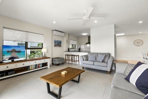 Una sala de estar moderna con televisor, sofá, mesa de centro y una cocina al fondo.