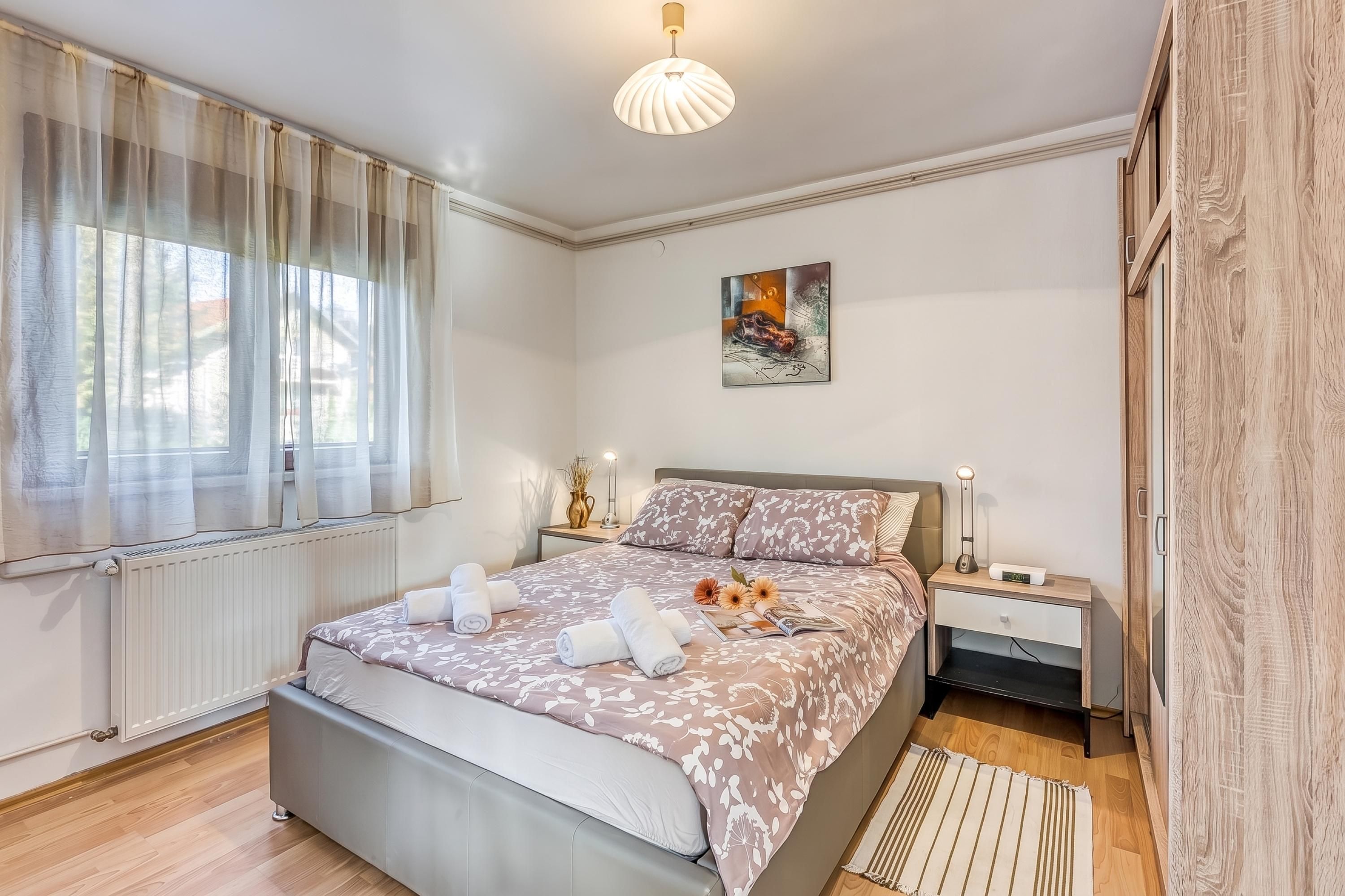 A modern bedroom with a large bed, a bedside table with a lamp, a radiator, and a window with sheer curtains.