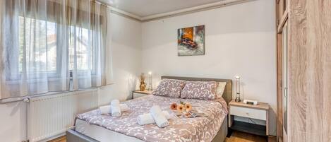 A modern bedroom with a large bed, a bedside table with a lamp, a radiator, and a window with sheer curtains.
