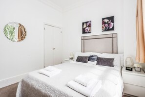 A neatly made bed with white linens and a fluffy blanket, a bedside table with a lamp, and a wall-mounted mirror.