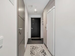 Un couloir étroit, avec une porte noire au fond, des murs blancs et un tapis à motifs sur le sol.