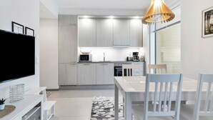 Een moderne keuken met witte kasten, een magnetron en een koffiezetapparaat. Er is een eettafel met stoelen en een aan de muur bevestigde televisie.
