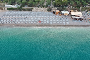 An aerial view of a beach with rows of sun loungers and umbrellas, a small building, and a pathway leading to it.