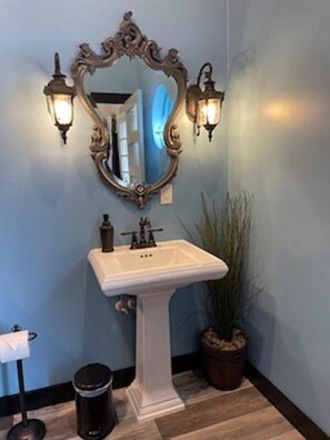 A bathroom with a white pedestal sink, ornate mirror, and wall-mounted sconces. - Tiny House Near Downtown Astoria (Astoria)