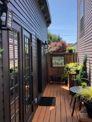 A wooden deck with a bench and potted plants, a wooden fence, and a building with large windows and a door. - Tiny House Near Downtown Astoria (Astoria)