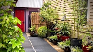 A narrow pathway flanked by potted plants and a wooden fence, leading to a red door. - Tiny House Near Downtown Astoria (Astoria)