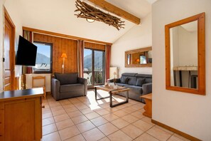 A living room with a sofa, a coffee table, a TV, and a large window with a view of mountains. - Appartement Courmayeur C44, Chamonix, France (Chamonix)