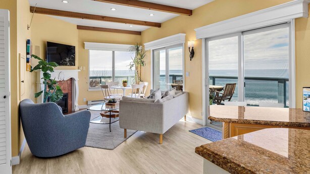 Een ruime woonkamer met een groot raam met uitzicht op de oceaan, een open haard en een eetgedeelte met panoramisch uitzicht.