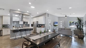 A modern kitchen with white cabinets, a dining area with a wooden table and benches, and a living room with a sofa and a TV.