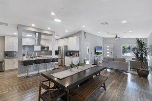 A modern kitchen with white cabinets, a dining area with a wooden table and benches, and a living room with a sofa and a TV.