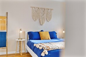 Une chambre à coucher avec un lit, des lampes de chevet, une décoration murale en macramé et une petite table avec une lampe.