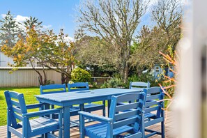 Une table et des chaises bleues, placées à l’extérieur sur une terrasse en bois, offrent une vue sur un jardin et des arbres.