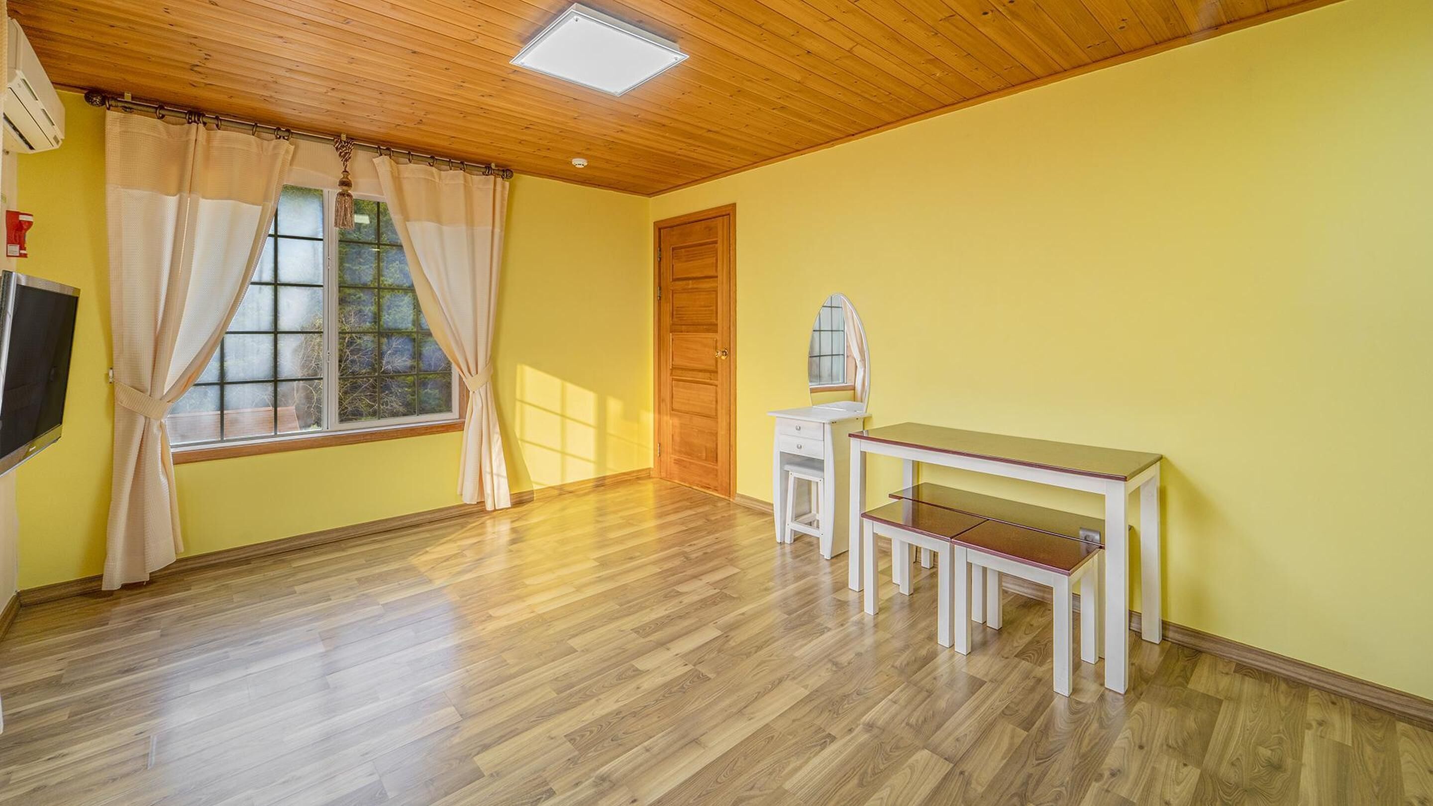 A room with wooden flooring, a wooden ceiling, a window with curtains, a door, and a small white desk with a mirror.