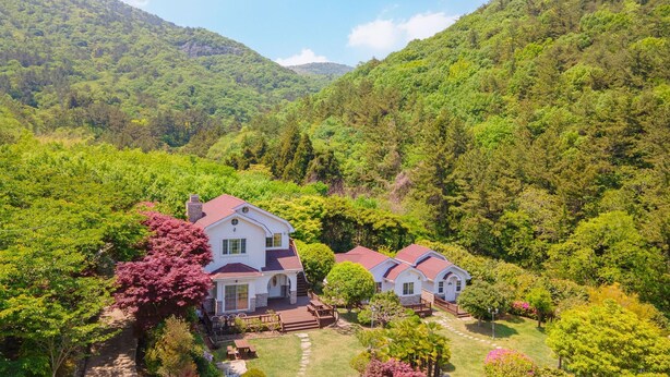 Una casa grande de color blanco con techo rojo rodeada de abundante vegetación y colinas.