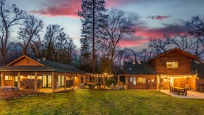 A log cabin with a porch and outdoor seating area, surrounded by trees and a sunset sky. - Log Cabin sleeps 20 with hot tub, EV Charger, firepit (Ahwahnee)