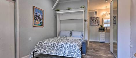 Un dormitorio con una cama, un ventilador de techo, un cuadro y una cocina al fondo.