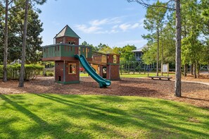 Un parque infantil con tobogán y estructuras para escalar, rodeado de árboles y un espacio cubierto de césped.