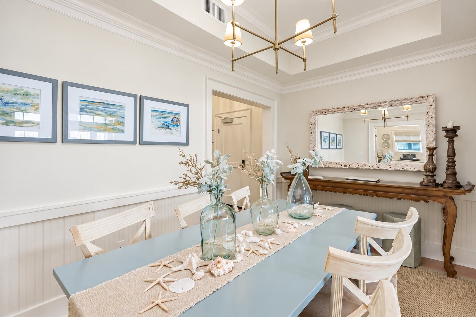 A dining area with a long table, chairs, framed artwork on the wall, and a mirror above the table.