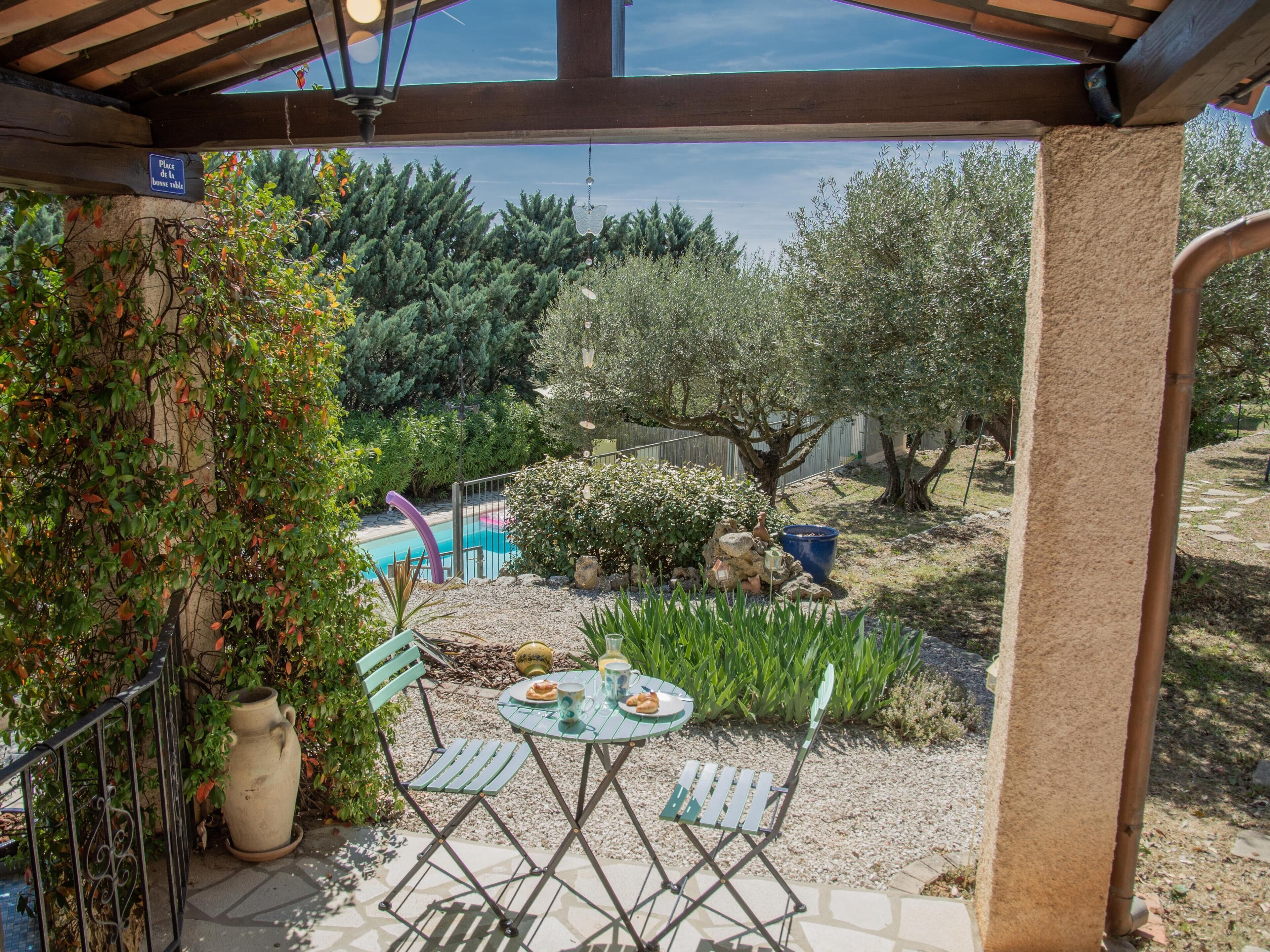 An outdoor patio area with a table and chairs, surrounded by greenery and a pool.