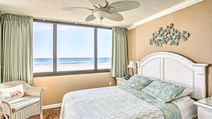 Una camera da letto con un'ampia finestra vista spiaggia, un letto con copriletto decorato, una sedia in vimini e una ventola a soffitto.