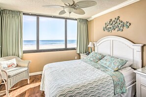 Un dormitorio con un amplio ventanal con vista a la playa, una cama con colcha estampada, una silla de mimbre y un ventilador de techo.