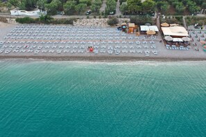 Une vue aérienne d’une plage avec des rangées de chaises longues et de parasols, un petit bâtiment et un chemin menant à celui-ci.
